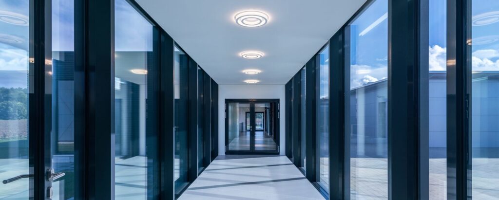 Stilrena cirkulära takarmaturer från PROLJUS AB i en ljus glasad korridor, energieffektiva LED‑armaturer som ger mjukt, jämnt ljus och förhöjer moderna kontors‑ och offentliga miljöer