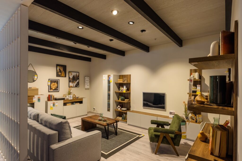 Vardagsrumsshowroom belyst av svarta takbalkar med infällda runda LED‑downlights och riktade spotlights som ger varm, jämn accentbelysning över möbler och väggar, energieffektiva armaturer i modern design perfekta för butik och hem, kontakta PROLJUS AB för belysningslösningar och beställning