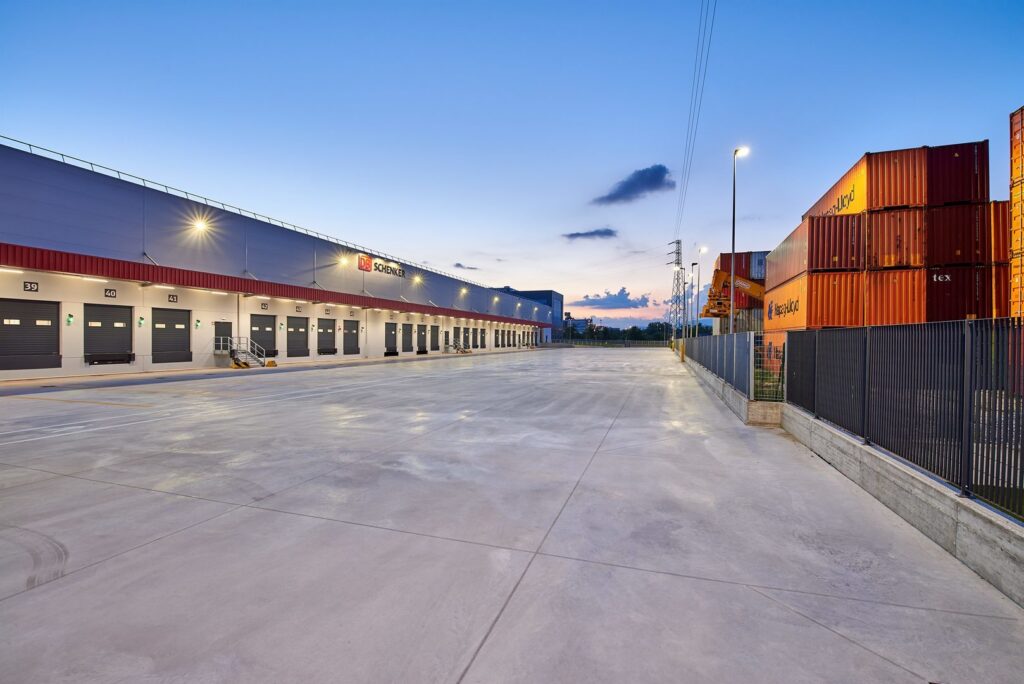 Skymningsbild av lager och containerområde där PROLJUS AB:s energieffektiva armaturer längs takfoten och på belysningsstolpar ger jämnt starkt ljus över lastkajer och uppfart för ökad säkerhet, driftseffektivitet och lägre elkostnader för logistikföretag