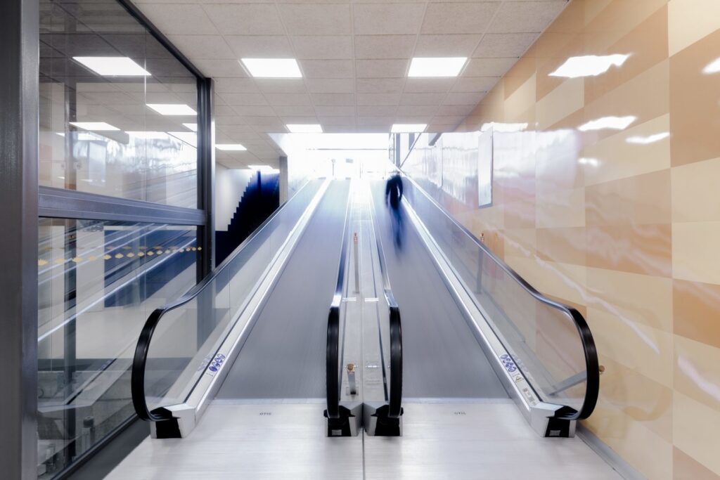 Kvadratiska infällda LED-armaturer i taket sprider jämnt, bländfritt ljus över en modern rulltrappa och reflekteras i glas och glänsande väggytor, ett energieffektivt och stilrent belysningskoncept från PROLJUS AB