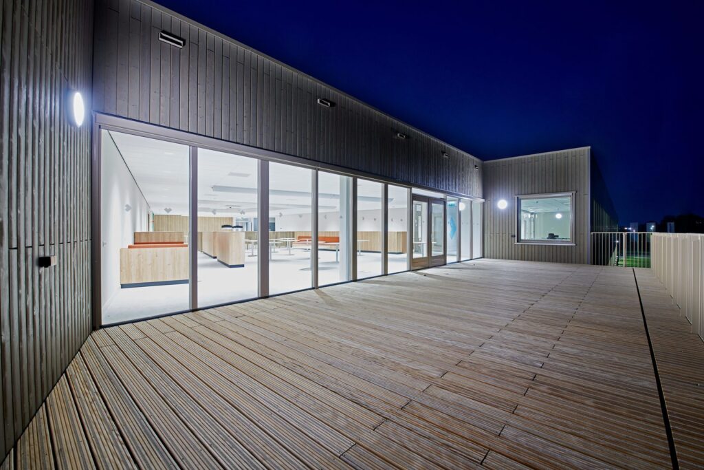 Nattbild av modern glasfasad och trädäck upplyst av utomhusarmaturer från PROLJUS AB med runda vägglampor och diskreta infällda LED-armaturer som ger jämnt, energieffektivt och tryggt ljus som framhäver arkitekturen och passar kommersiella entréer