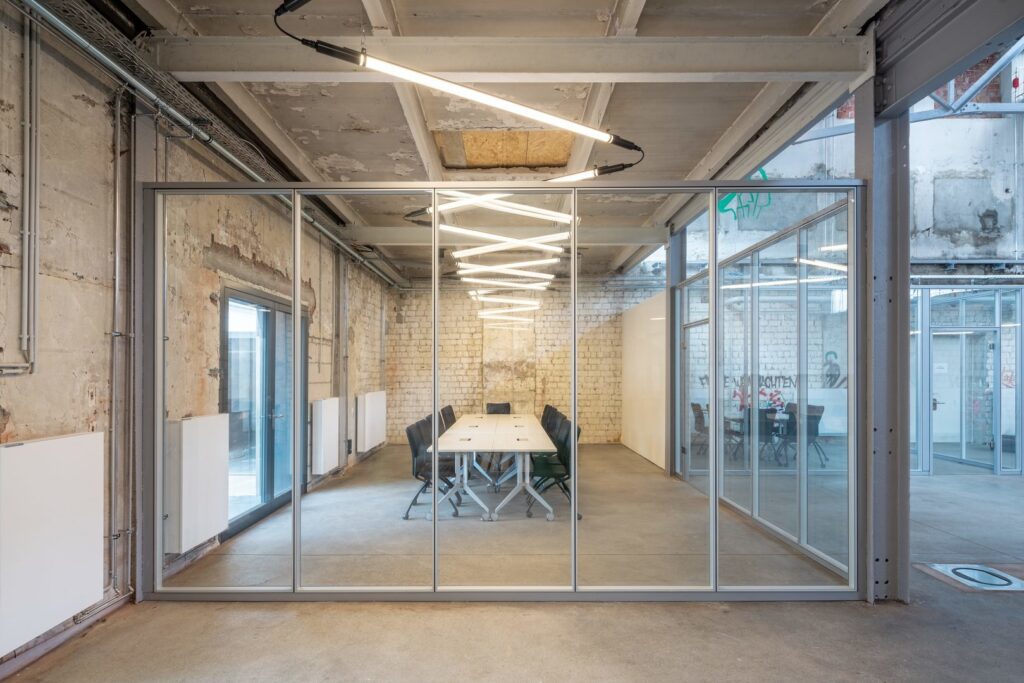 Zigzagformade LED-luminärer i varmvitt ljus hänger över ett glasat konferensrum i industriell kontorsmiljö, elegant och energieffektiv belysning från PROLJUS AB som förhöjer arbetsmiljön