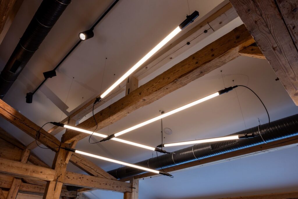 Takinstallation med synliga träbalkar och flera långa varmvita linjära LED-armaturer hängande i geometriskt mönster med svarta kablar och fästen, industriell och elegant atmosfär perfekt för kontor eller restauranger levererade av PROLJUS AB med energieffektiv modern design
