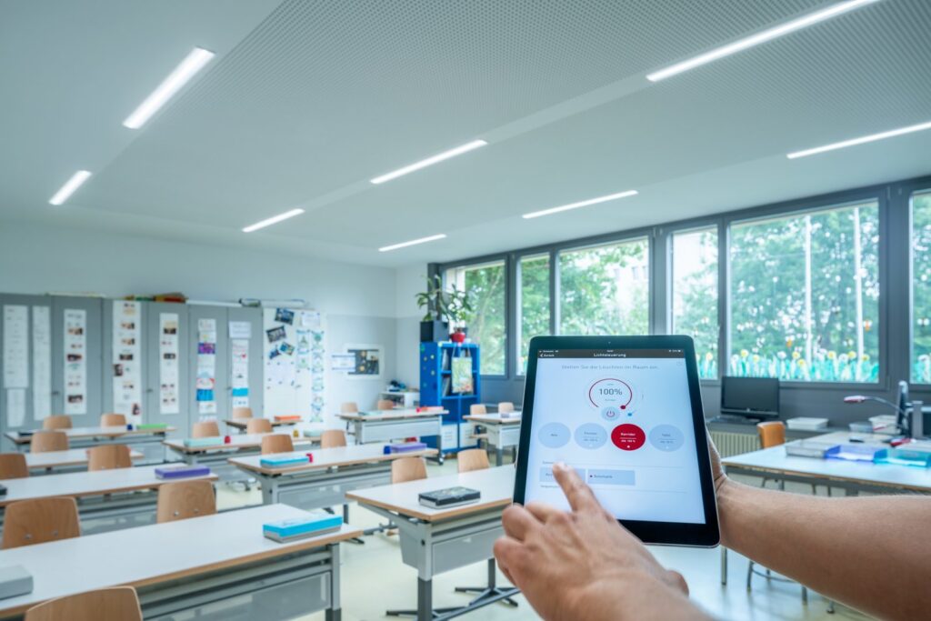 Modernt klassrum med jämnt fördelade slanka LED-armaturer i taket från PROLJUS AB, dimbara och energieffektiva, styrda via surfplatta i förgrunden, idealiska för skolor och arbetsmiljöer