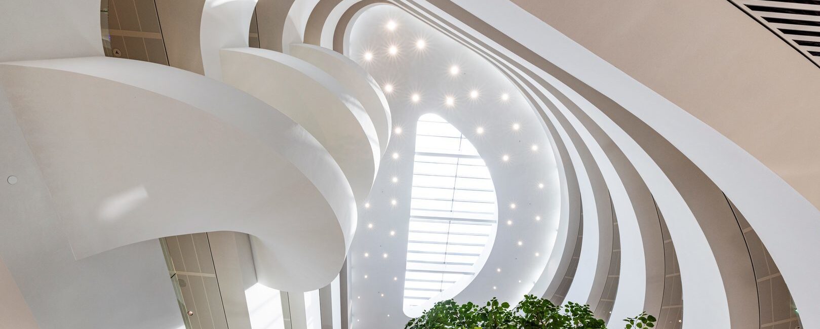 Modernt atrium med böljande balkonger, stort takfönster och grönska – futuristisk kontorsarkitektur inomhus.
