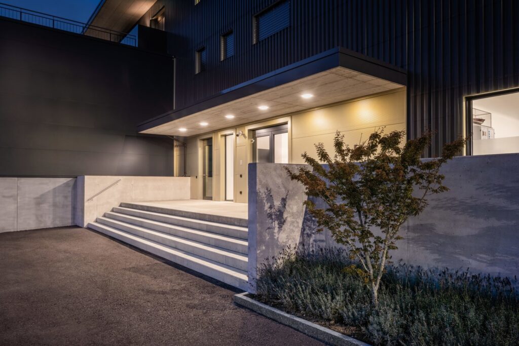 Kvällsentré upplyst av PROLJUS AB:s infällda varmvita LED-armaturer som jämnt belyser trappan, betongväggarna och växterna och skapar en trygg, elegant och energieffektiv entrélösning, kontakta PROLJUS AB för offert