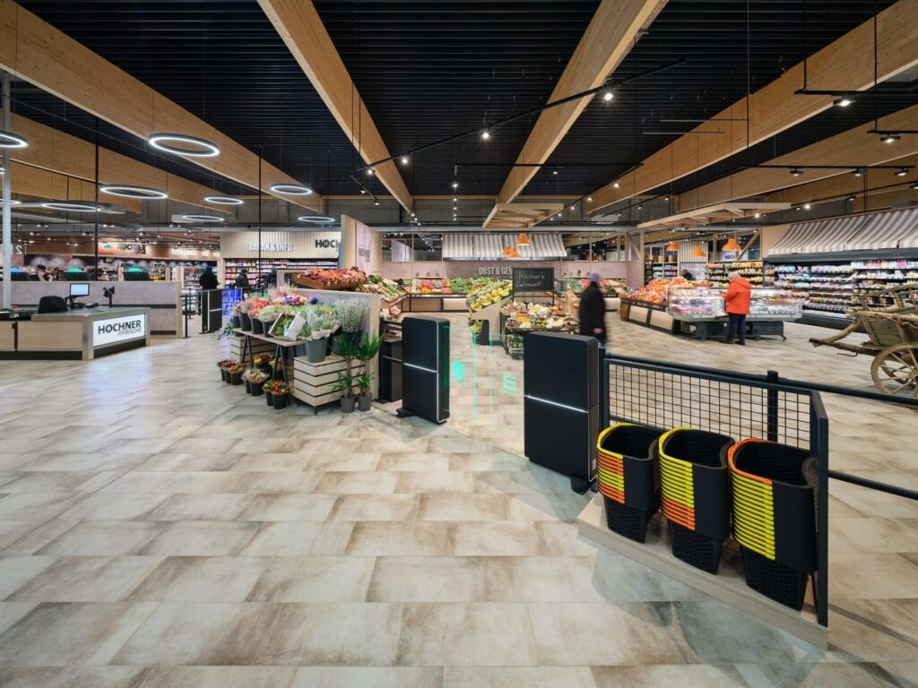 Stort modernt butiksgolv upplyst av eleganta cirkulära ringarmaturer och linjära spottar monterade i takets svarta lameller och varma träbalkar, energieffektiv LED-belysning som framhäver frukt och blommor och skapar en inbjudande atmosfär perfekt för butikslösningar från PROLJUS AB som ökar synlighet och försäljning