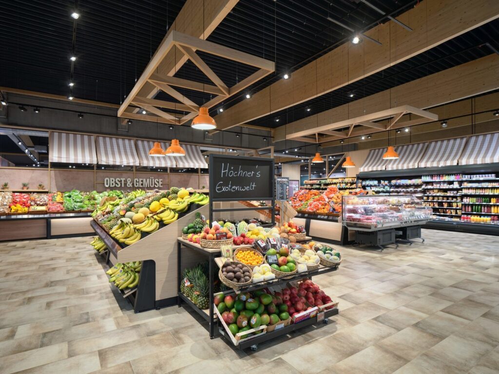 Stilren frukt- och grönsaksavdelning i ett matvaruhus med prydliga displayer fyllda med bananer, mango, ananas och citrus, hängande orange lampor och synliga träramar i taket samt skyltar med OBST & GEMÜSE och en griffeltavla där det står Höchners Exotenwelt