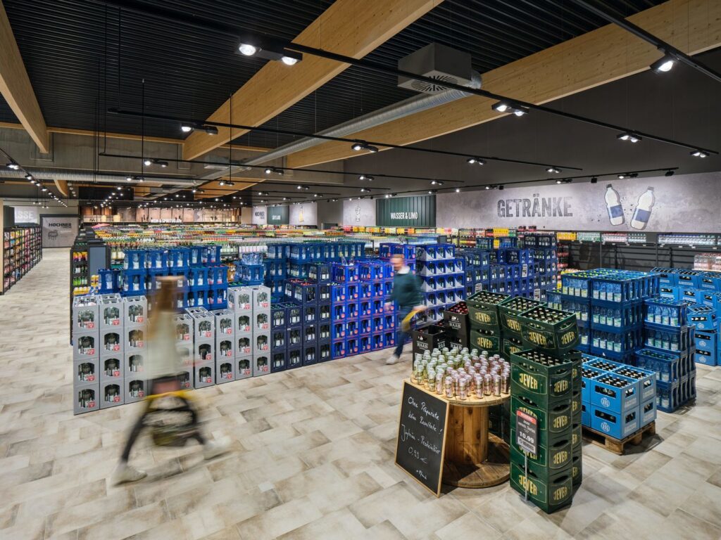 Stor livsmedelsbutiks dryckesavdelning sedd snett uppifrån med staplade öl‑ och vattenbackar i blått och grönt, ett runt bord med burkar i förgrunden, breda gångar och två suddiga kunder som rör sig med kundvagn under högt tak med synliga träbjälkar och belysning