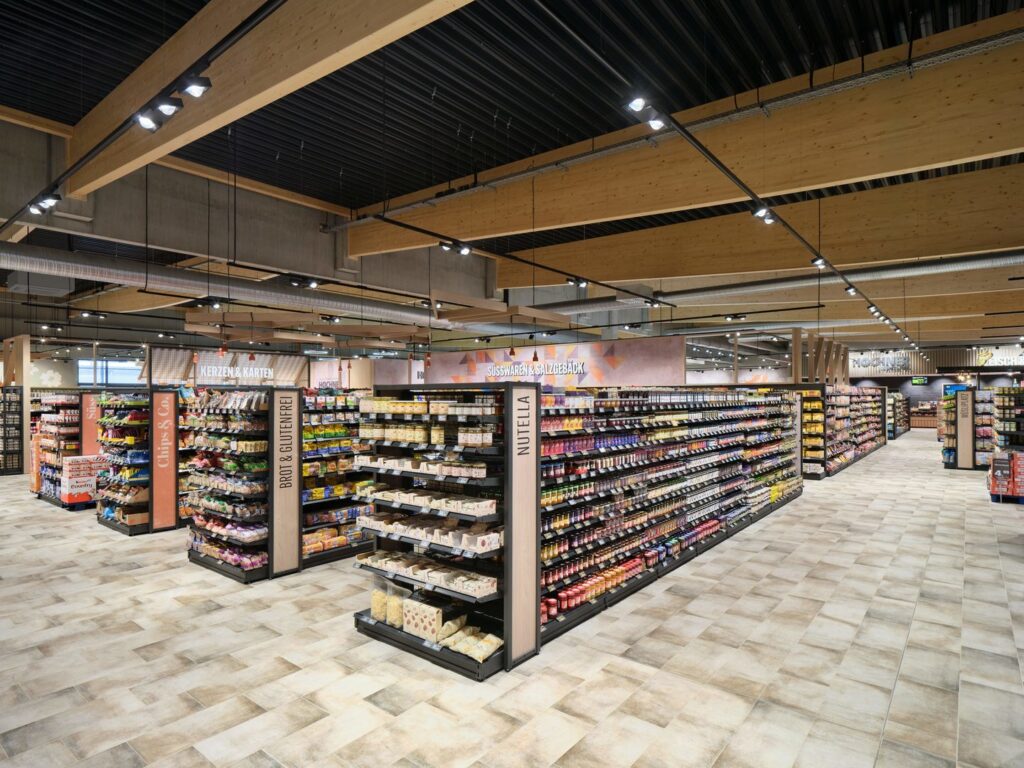 Modern och välorganiserad livsmedelsbutik med hyllor fyllda med olika produkter, ljusa golv och trädetaljer i taket.