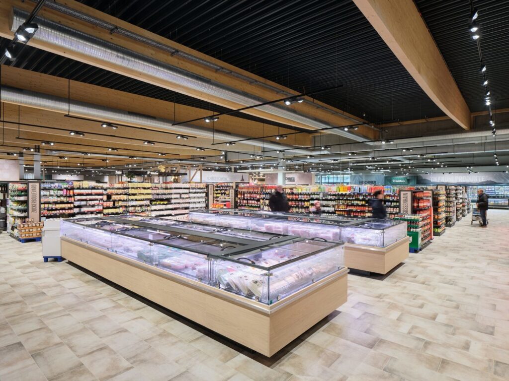 Stor ljus livsmedelsbutik med breda gångar och låga glasförsedda frysdiskar i mitten, väl upplyst tak med synliga ventilationskanaler och träbalkar, hyllor fyllda med varor och några kunder